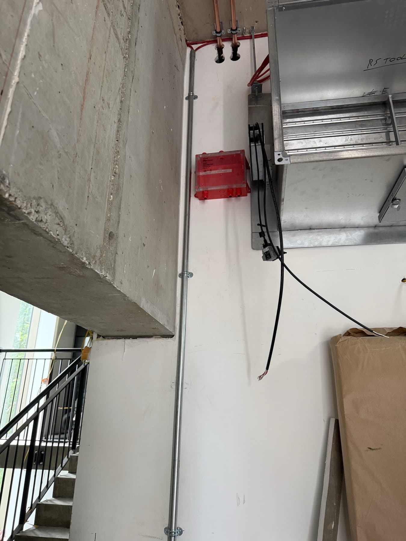 Galvanised steel conduit clipped up a stairwell wall beside a concrete beam, next to a red surface-mounted connection box and galvanised ductwork