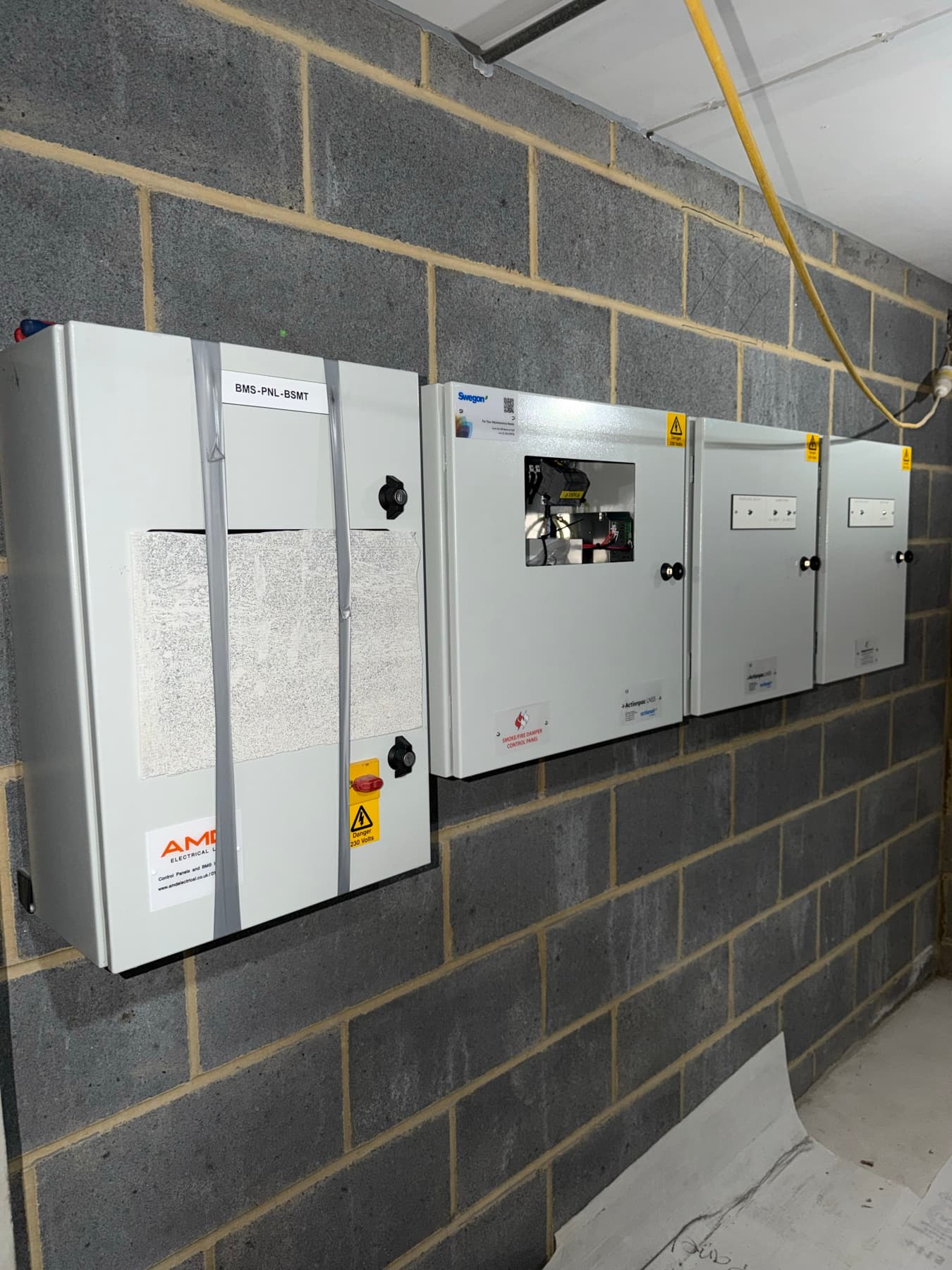 Four grey control panels wall-mounted on a blockwork wall: a BMS panel labelled BMS-PNL-BSMT with banding straps and protective sheeting still on its door, beside three smaller panels, one labelled as a Swegon smoke/fire damper control panel