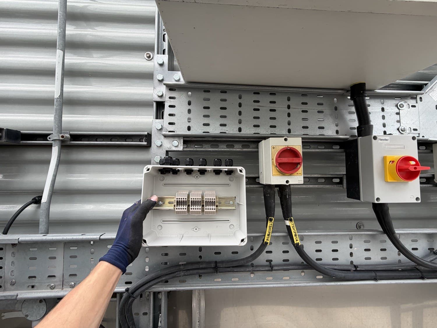 Gloved hand at an open terminal junction box with DIN-rail terminals, beside rotary isolators and galvanised cable containment