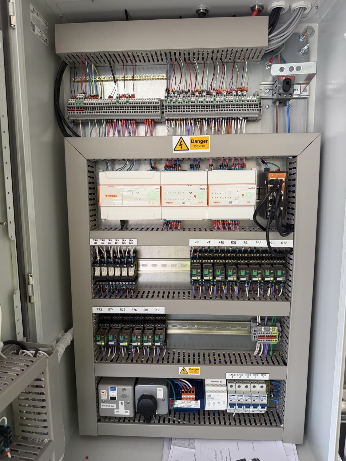 Interior of a control panel with a Trend IQ500 outstation and input/output modules, a network switch, rows of labelled interface relays and labelled terminal blocks